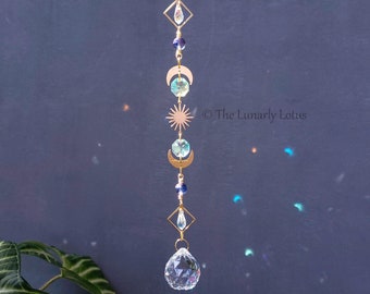 May include: A crystal suncatcher with a gold chain and a large clear crystal at the bottom. The suncatcher features a series of gold crescent moons, a gold sun, and a gold diamond shape with a small crystal inside. The text "The Lunarly Lotus" is visible in the image.