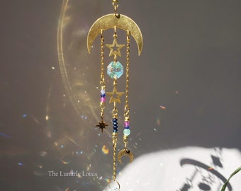 May include: A gold crescent moon with stars and crystals hanging from it. The crystals are iridescent and reflect light. The moon and stars are attached to a chain that ends with a large iridescent crystal. The text "The Lunarly Lotus" is visible at the bottom of the image.