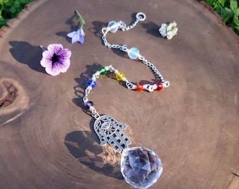 Puede incluir: Un atrapasol de cristal con un amuleto de mano de hamsa plateado y cuentas de colores arcoíris que cuelgan de una cadena plateada. El atrapasol está colgado de una rodaja de madera.