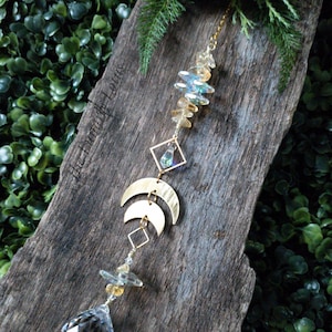 May include: A crystal suncatcher with a gold chain, featuring a crystal prism, gold crescent moons, and a cluster of iridescent crystals. The suncatcher is hanging from a wooden board with a green leafy background.