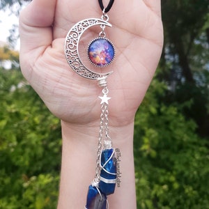 Puede incluir: Un colgante de media luna de plata con un cabuj&oacute;n de vidrio con tem&aacute;tica de galaxia, que cuelga de un cord&oacute;n negro. Un amuleto de estrella de plata cuelga de la media luna, con dos puntas de cristal azul envueltas en alambre de plata que cuelgan debajo.