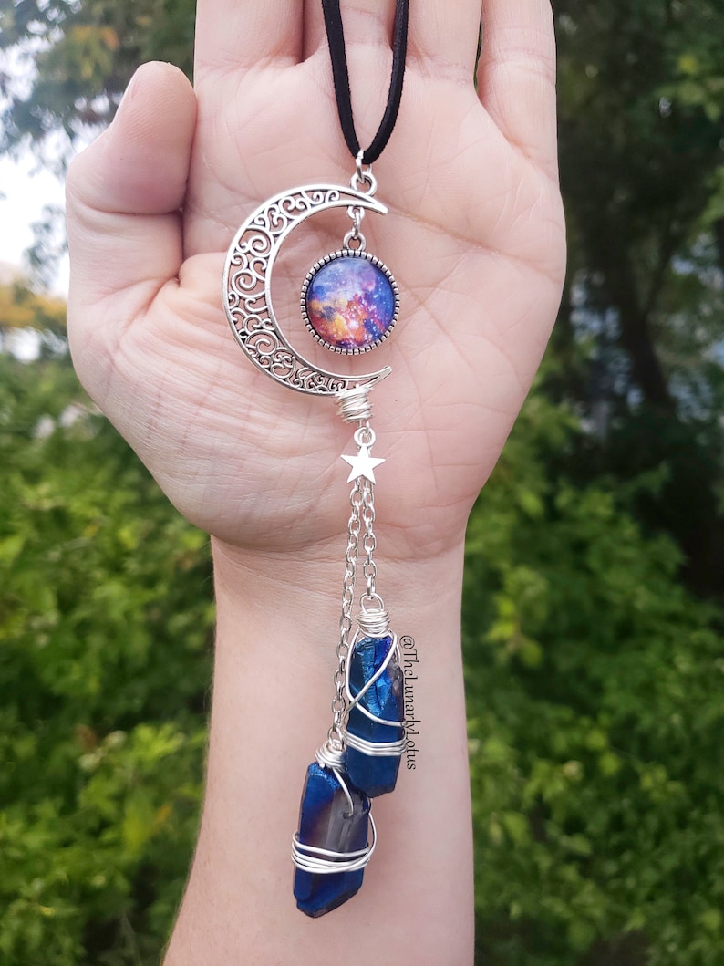Puede incluir: Un colgante de media luna de plata con un cabuj&oacute;n de vidrio con tem&aacute;tica de galaxia, que cuelga de un cord&oacute;n negro. Un amuleto de estrella de plata cuelga de la media luna, con dos puntas de cristal azul envueltas en alambre de plata que cuelgan debajo.