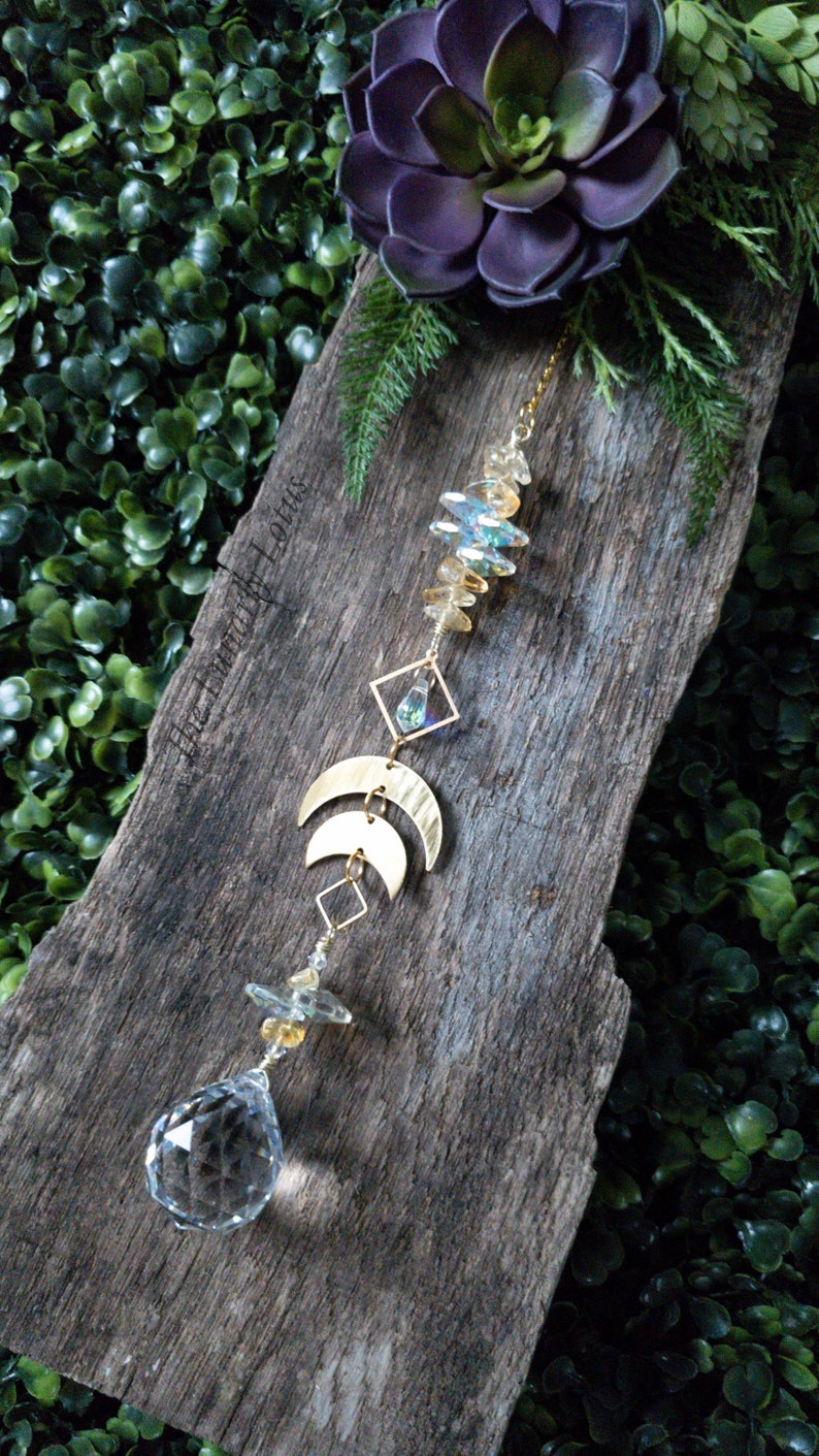 May include: A crystal suncatcher with gold accents and a clear crystal ball hanging from a wooden board. The suncatcher features a geometric design with crescent moons and a small crystal in a square frame. The board is surrounded by green foliage.