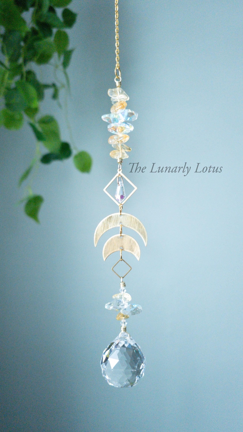May include: A gold crystal suncatcher with a crystal ball at the bottom. The suncatcher features a gold chain, a cluster of clear and gold crystals, a gold diamond shape, and two gold crescent moons. The text "The Lunarly Lotus" is visible in the image.