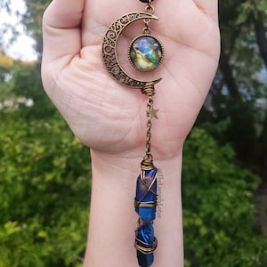 Puede incluir: Un colgante de media luna dorado con un cabuj&oacute;n de vidrio con tem&aacute;tica de galaxia y una punta de cristal azul envuelta en alambre. El colgante cuelga de un cord&oacute;n negro.
