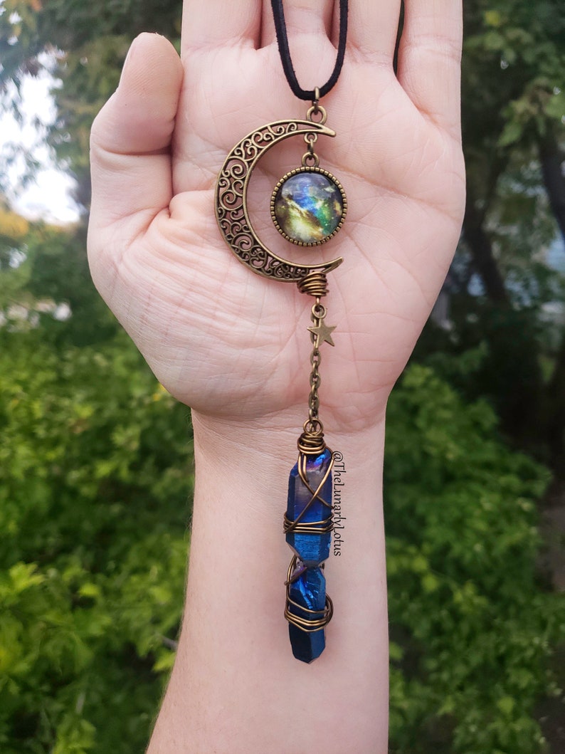 Puede incluir: Un colgante de media luna dorado con un cabuj&oacute;n de vidrio con tem&aacute;tica de galaxia y una punta de cristal azul envuelta en alambre. El colgante cuelga de un cord&oacute;n negro.
