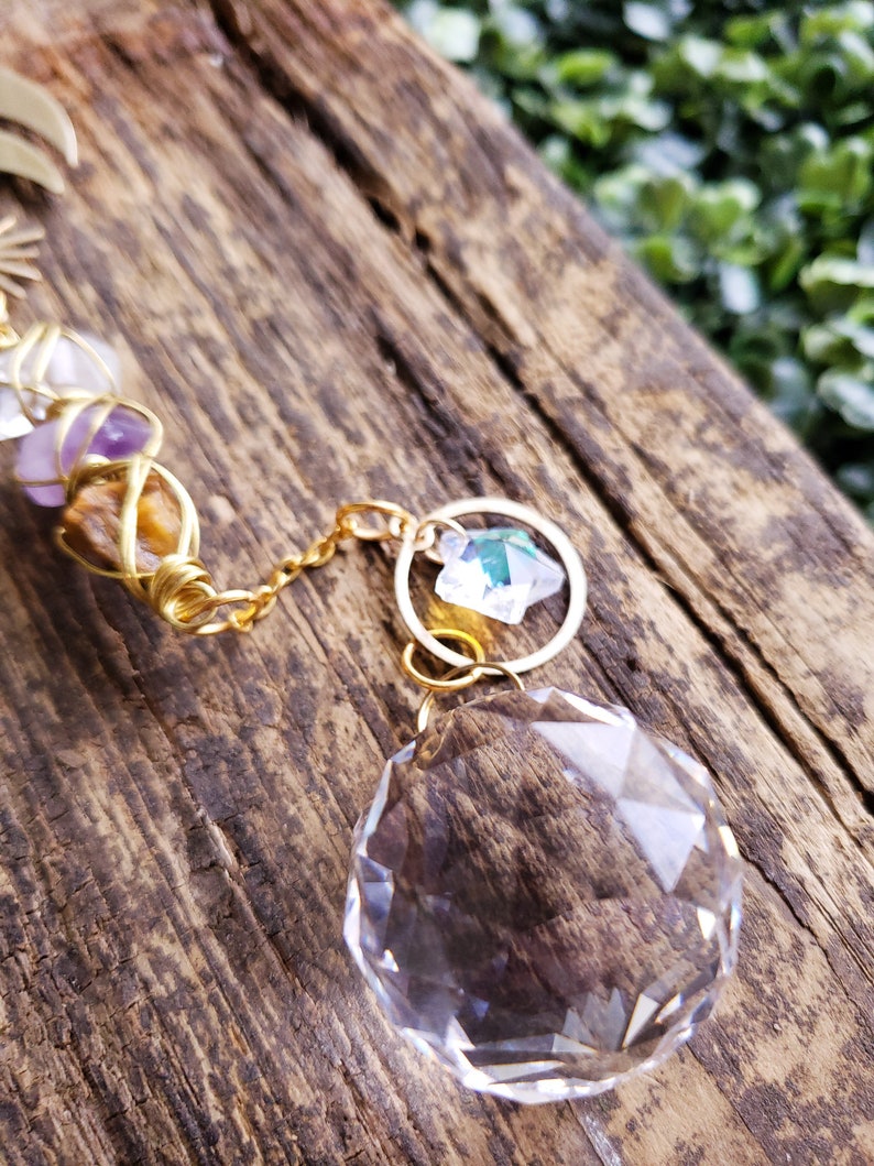 K&ouml;nnte beinhalten: Eine goldene Kette mit einer Kristallkugel und einem kleineren Kristallanh&auml;nger, die daran h&auml;ngen. Die Kette ist um ein St&uuml;ck Holz gewickelt.
