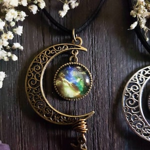 Puede incluir: Un collar con un colgante de media luna con un cabuj&oacute;n de vidrio con tem&aacute;tica de galaxia. El colgante est&aacute; hecho de bronce y tiene un delicado dise&ntilde;o de filigrana. El collar est&aacute; en un cord&oacute;n negro.