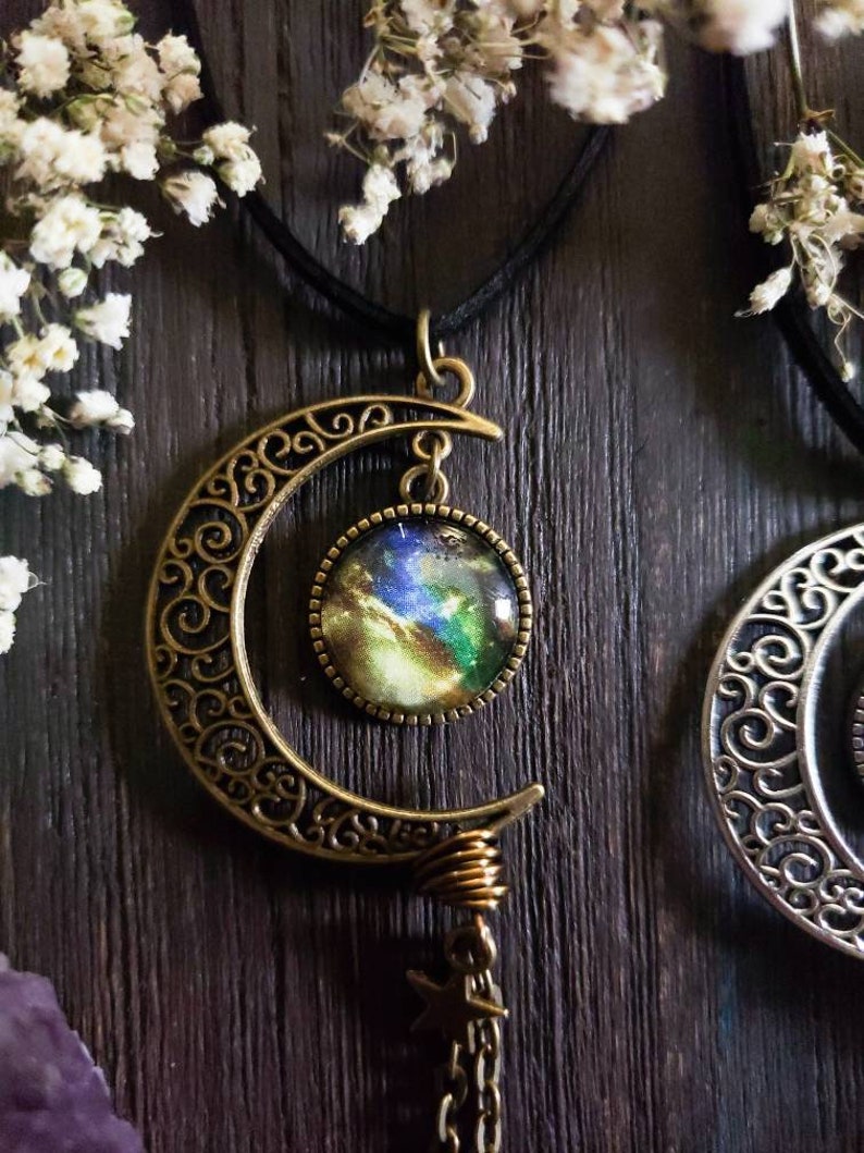 Puede incluir: Un collar con un colgante de media luna con un cabuj&oacute;n de vidrio con tem&aacute;tica de galaxia. El colgante est&aacute; hecho de bronce y tiene un delicado dise&ntilde;o de filigrana. El collar est&aacute; en un cord&oacute;n negro.
