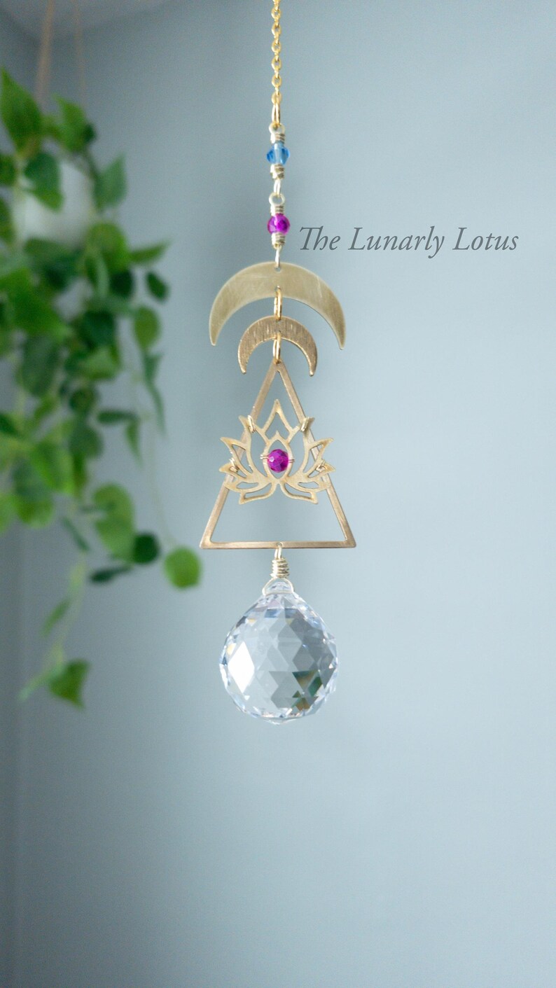 K&ouml;nnte beinhalten: Ein goldfarbener H&auml;ngeschmuck mit einem Kristallprisma, einem Lotusblumen-Design und drei Halbmonden. Der Anh&auml;nger ist an einer Kette mit kleinen blauen und rosa Perlen befestigt. Der Text "The Lunarly Lotus" ist auf dem Bild zu sehen.