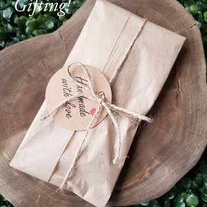 K&ouml;nnte beinhalten: Ein braunes Papierpaket, das mit Schnur gebunden ist und einen runden Anh&auml;nger mit der Aufschrift "Handgefertigt mit Liebe" tr&auml;gt. Das Paket liegt auf einer Holzoberfl&auml;che.