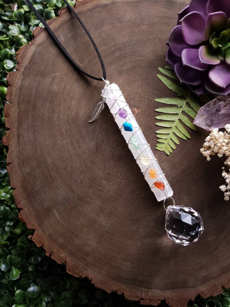 Puede incluir: Una varita de cristal blanco con siete piedras de chakra envueltas en alambre, un amuleto de pluma de plata y un colgante de cristal transparente que cuelga de un cord&oacute;n negro.