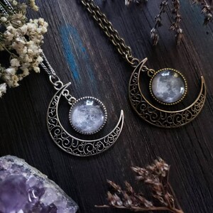 May include: Two silver crescent moon necklaces with glass moon pendants. The necklaces are on a dark wooden surface with dried flowers and a purple amethyst crystal.