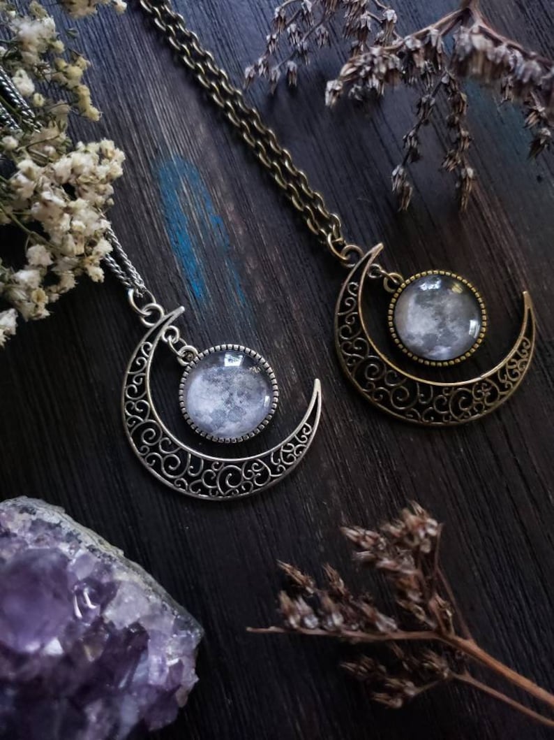 May include: Two silver crescent moon necklaces with glass moon pendants. The necklaces are on a dark wooden surface with dried flowers and a purple amethyst crystal.