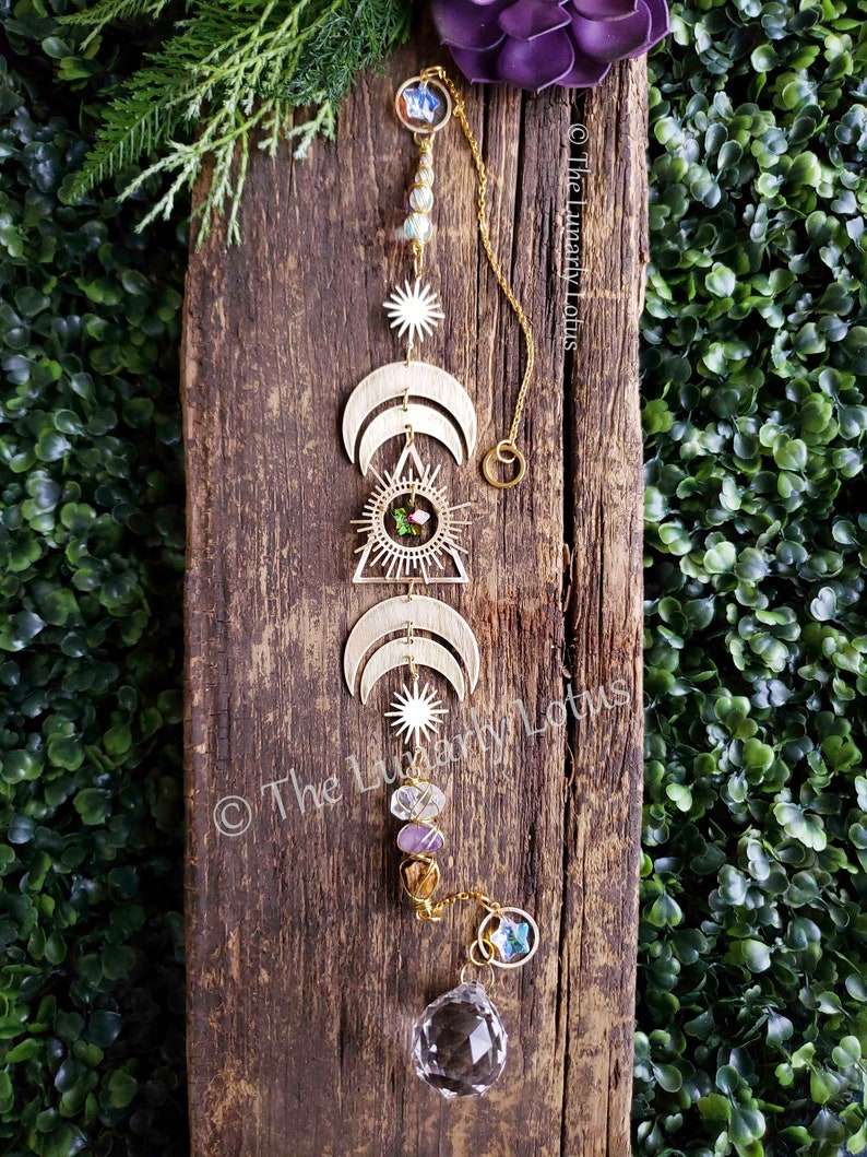 K&ouml;nnte beinhalten: Goldfarbener Metall-Sonnenf&auml;nger mit einem Kristallprisma, drei Mondsicheln, einem Dreieck mit einem Sonnenstrahl und einer Perlenkette mit Amethyst und klaren Quarzkristallen. Der Sonnenf&auml;nger h&auml;ngt an einer Holzoberfl&auml;che.