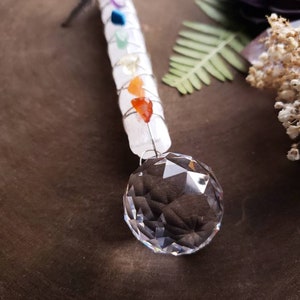 Puede incluir: Una varita de selenita blanca con siete piedras de chakra envueltas alrededor, coronada con una bola de cristal transparente. La varita cuelga de un cord&oacute;n negro.
