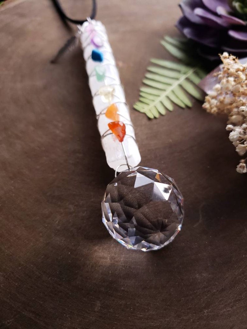 Puede incluir: Una varita de selenita blanca con siete piedras de chakra envueltas alrededor, coronada con una bola de cristal transparente. La varita cuelga de un cord&oacute;n negro.