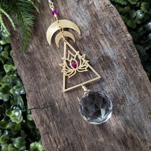 K&ouml;nnte beinhalten: Goldfarbener, h&auml;ngender Metallschmuck mit einem Kristallprisma, einem Lotusbl&uuml;ten-Design und drei Halbmonden. Der Schmuck h&auml;ngt an einer Kette mit Perlen.