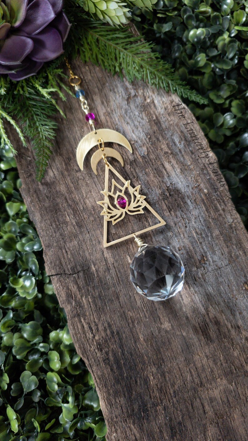 K&ouml;nnte beinhalten: Goldfarbener, h&auml;ngender Metallschmuck mit einem Kristallprisma, einem Lotusbl&uuml;ten-Design und drei Halbmonden. Der Schmuck h&auml;ngt an einer Kette mit Perlen.