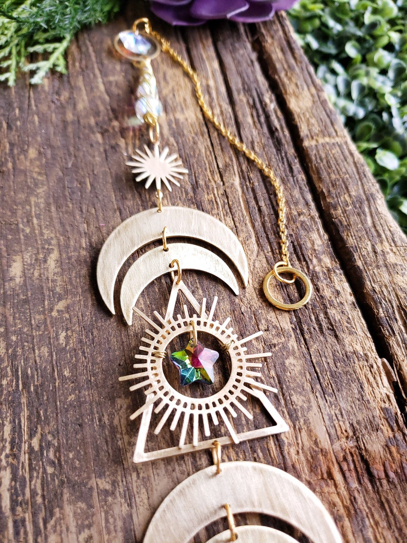K&ouml;nnte beinhalten: Ein goldfarbener Metall-Sonnen- und Mond-Mobile mit einem Kristallstern, der in der Mitte h&auml;ngt. Das Mobile h&auml;ngt an einer goldenen Kette und ist an einer Holzoberfl&auml;che befestigt.