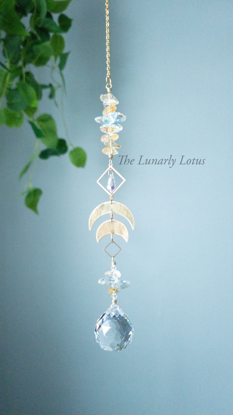 May include: A gold crystal suncatcher with a crystal ball at the bottom. The suncatcher features a geometric design with two crescent moons and a crystal in a square frame. The text "The Lunarly Lotus" is visible in the image.