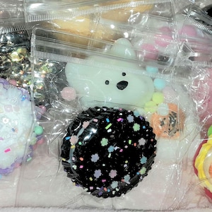 May include: Assortment of colourful, squishy, and decorative charms in various shapes, including a cake, a donut, a sandwich, a cloud, and a star. The charms are packaged in clear plastic bags.