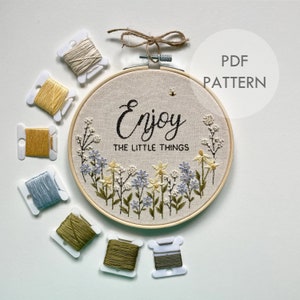 Pode incluir: Um aro de bordado de madeira com um fundo de tecido bege que apresenta um design floral e o texto "Enjoy the little things". O aro é rodeado por oito pequenas bobinas de fio de bordar de várias cores.