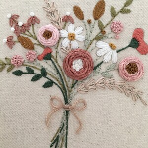以下が含まれることがあります： ピンクと白の花のブーケを手刺繍した作品です。緑の葉と茎があり、茶色のリボンで結ばれています。