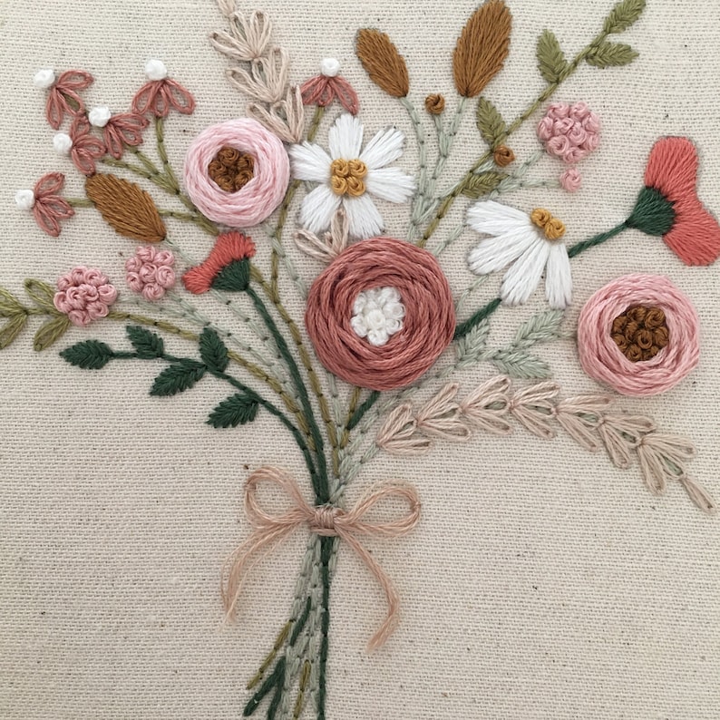 以下が含まれることがあります： ピンクと白の花のブーケを手刺繍した作品です。緑の葉と茎があり、茶色のリボンで結ばれています。