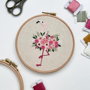 May include: Embroidered hoop art featuring a pink flamingo with a floral bouquet made of pink and white flowers. The hoop is surrounded by spools of embroidery floss in various shades of pink, green, and white.