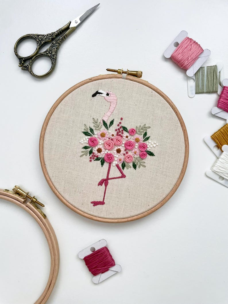 May include: Embroidered hoop art featuring a pink flamingo with a floral bouquet made of pink and white flowers. The hoop is surrounded by spools of embroidery floss in various shades of pink, green, and white.