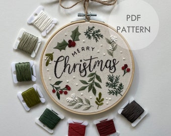 Puede incluir: Un aro de bordado blanco con fondo negro y el texto "Merry Christmas" en cursiva. El aro está rodeado de varios carretes de hilo de bordar de colores, incluyendo verde, rojo y marrón.
