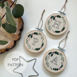 Puede incluir: Tres adornos navideños bordados con un diseño de corona de acebo y una letra en el centro. Las letras son H, P y A. Los adornos son blancos con bordados verdes y rojos.