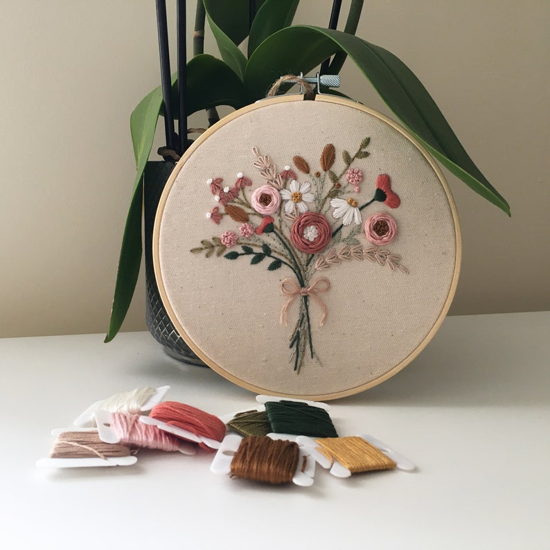 以下が含まれることがあります： ベージュの布地に手縫いのピンク、白、緑の花束が刺繍された木製刺繍枠。枠の周りは様々な色の刺繍糸で囲まれています。