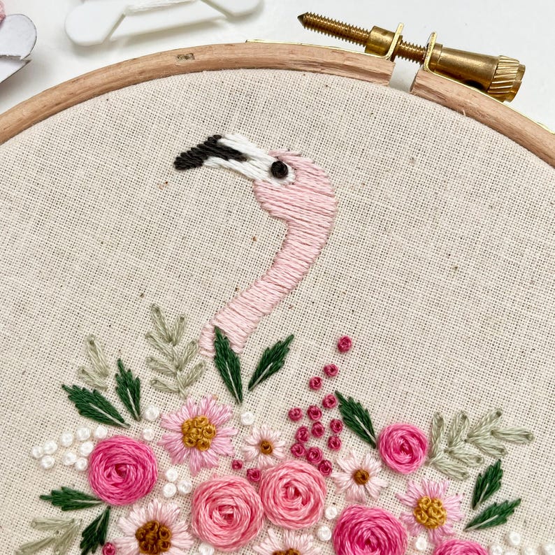 May include: Embroidered hoop art featuring a pink flamingo with a black beak and a floral arrangement of pink and white flowers with green leaves.