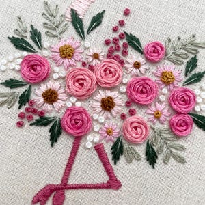 May include: Pink flamingo embroidered with pink and white flowers, green leaves, and small pink and white dots on a white fabric background.