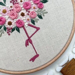 May include: Embroidered hoop art featuring a pink flamingo with a bouquet of pink and white flowers. The hoop is made of wood and has a natural finish.