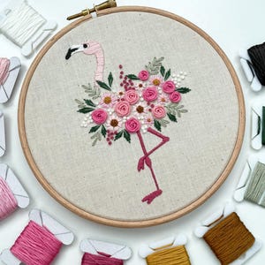 May include: Embroidered hoop art featuring a pink flamingo with a floral design. The flamingo is surrounded by pink, white, and green flowers. The hoop is made of wood and has a natural linen fabric.