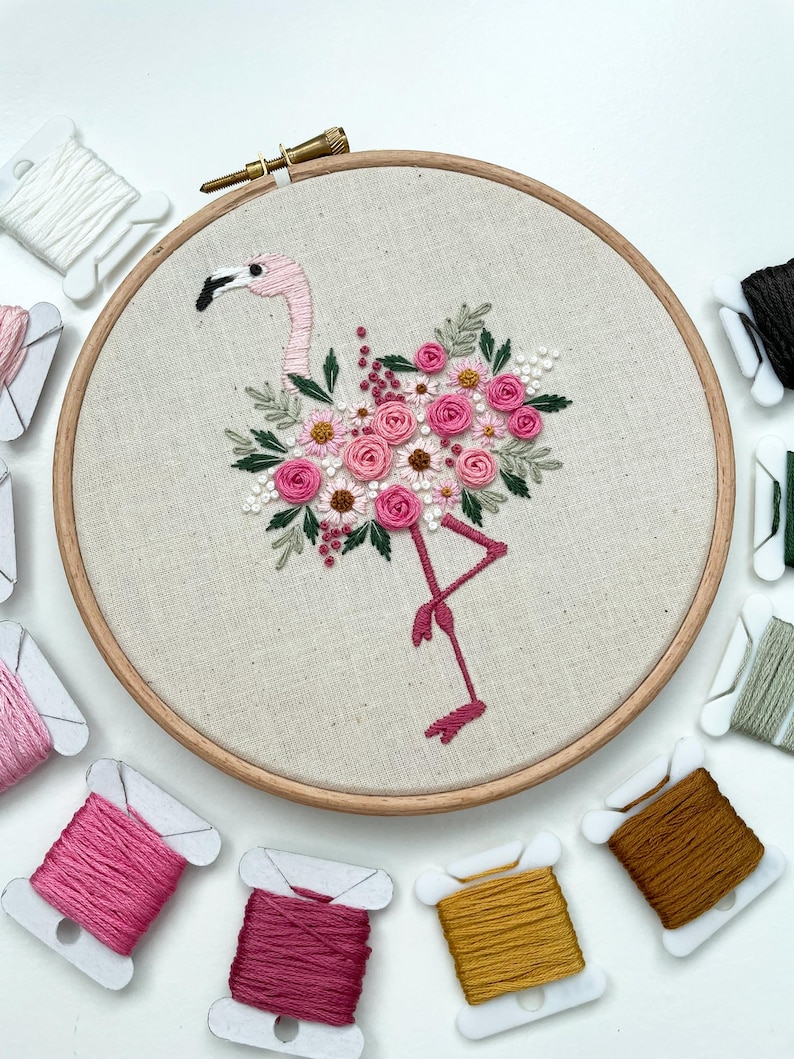 May include: Embroidered hoop art featuring a pink flamingo with a floral design. The flamingo is surrounded by pink, white, and green flowers. The hoop is made of wood and has a natural linen fabric.