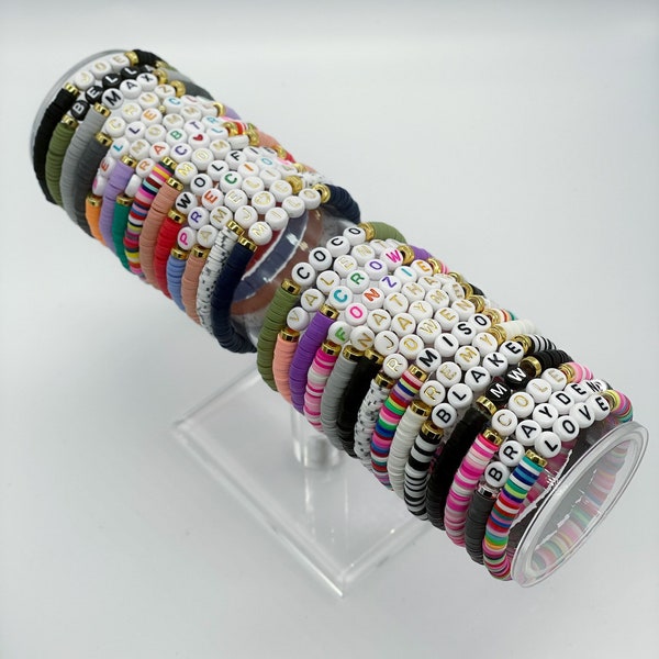 May include: A stack of colorful beaded bracelets with different names spelled out on them. The bracelets are arranged in a circle on a clear acrylic stand.