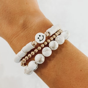 May include: A set of three bracelets featuring white howlite beads and gold spacers. One bracelet has a white smiley face charm.
