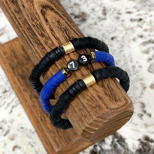 May include: Three bracelets stacked on a wooden display stand. The bracelets are made of black and blue beads with gold accents. The top bracelet has a black bead with the number "7" on it, the middle bracelet has a black bead with the number "3" on it, and the bottom bracelet is all black.