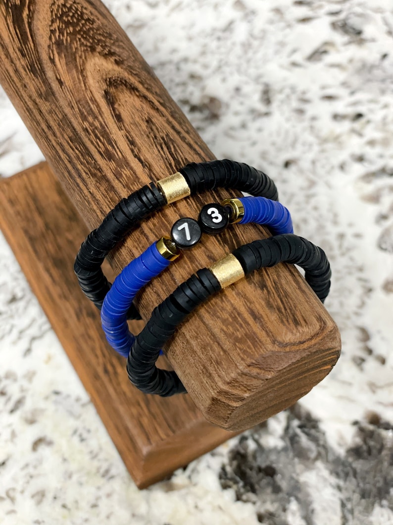 May include: Three bracelets stacked on a wooden display stand. The bracelets are made of black and blue beads with gold accents. The top bracelet has a black bead with the number "7" on it, the middle bracelet has a black bead with the number "3" on it, and the bottom bracelet is all black.