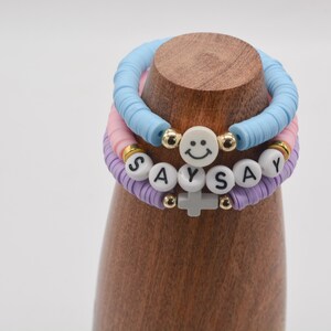 May include: Three colourful beaded bracelets with the word "SAY" spelled out on one bracelet and a smiley face charm on another. The bracelets are stacked on a wooden display stand.