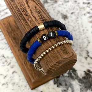 May include: Three bracelets stacked on a wooden display stand. The top bracelet is black with a gold accent. The middle bracelet is blue with white beads that spell out "97". The bottom bracelet is gold.