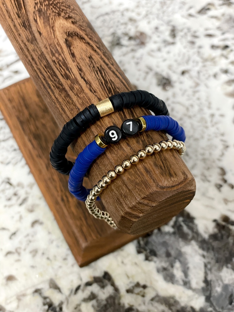 May include: Three bracelets stacked on a wooden display stand. The top bracelet is black with a gold accent. The middle bracelet is blue with white beads that spell out "97". The bottom bracelet is gold.