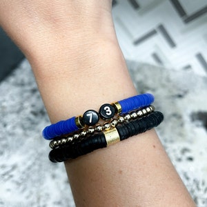 May include: Three bracelets stacked on a wrist. The top bracelet is blue and the bottom two are black. The top bracelet has two black beads with white numbers "7" and "3" on them.