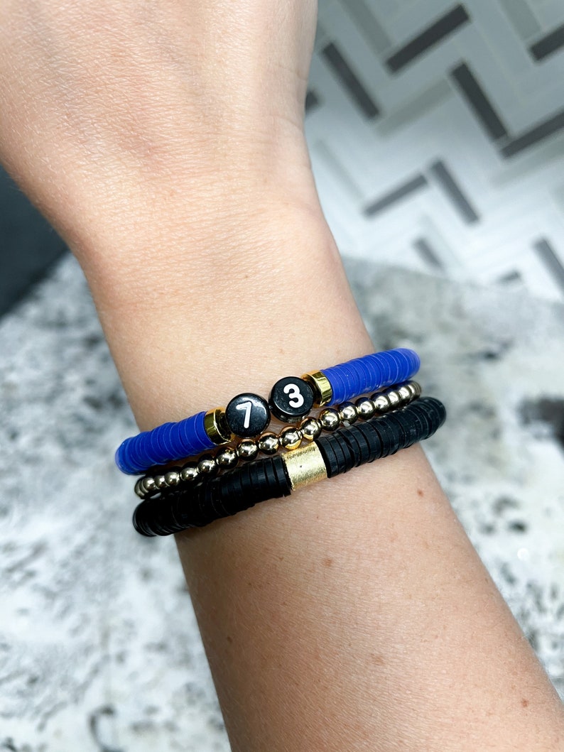 May include: Three bracelets stacked on a wrist. The top bracelet is blue and the bottom two are black. The top bracelet has two black beads with white numbers "7" and "3" on them.