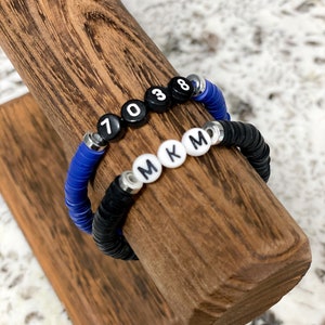 May include: Two bracelets on a wooden display stand. One bracelet is black with white letter beads that spell "MKM". The other bracelet is blue with black number beads that spell "7038".