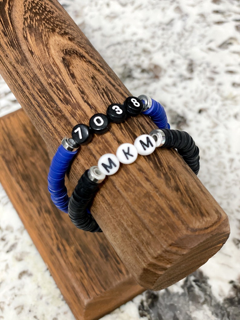 May include: Two bracelets on a wooden display stand. One bracelet is black with white letter beads that spell "MKM". The other bracelet is blue with black number beads that spell "7038".